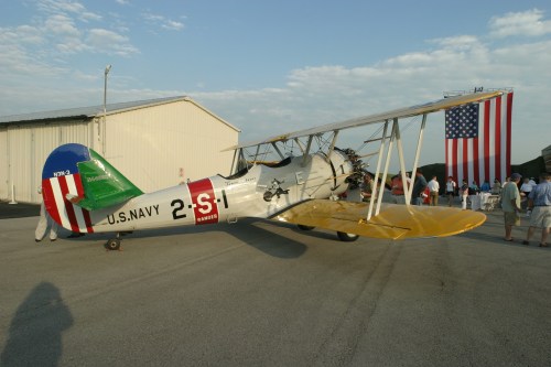 A vintage biplane with U.S. Navy markings parked near a hangar, with an American flag displayed in the background.