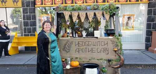 A woman in a green cloak stands by a decorated apothecary cart with plants, pumpkins, and Halloween-themed items.