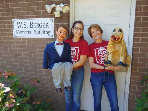 Two women stand outside a building, each holding a puppet—one a ventriloquist dummy and the other a dog figure.