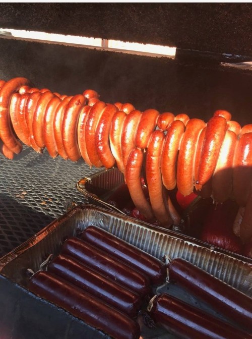 Grilled sausages hanging on a rack, with more sausages in a tray below, all in a smoky barbecue setting.