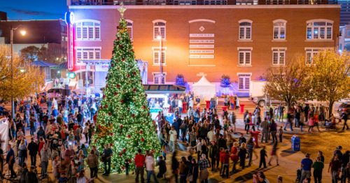 A festive crowd gathers around a large decorated Christmas tree in a lively downtown area at night.
