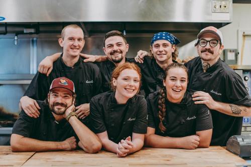 A group of seven chefs in black uniforms pose together in a kitchen, smiling and showing camaraderie.