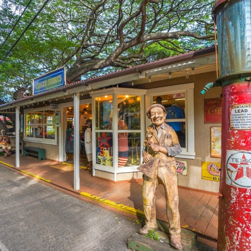 A wooden statue of a man stands outside a colorful shop with large windows and vintage gas pump nearby.