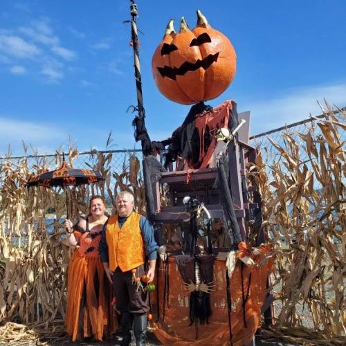 A couple in Halloween costumes stands beside a large pumpkin figure on a throne, surrounded by corn stalks.