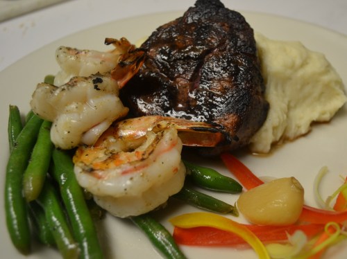 A plate featuring grilled shrimp, a charred steak, mashed potatoes, and green beans with colorful bell pepper strips.