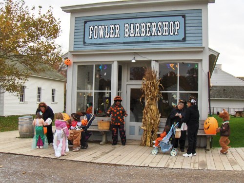 A barbershop decorated for Halloween, with children in costumes and adults outside, surrounded by autumn decor.