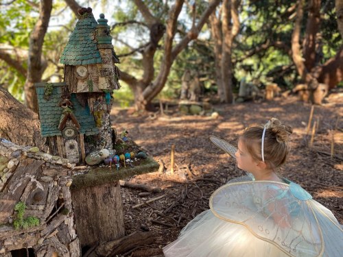 A child in a fairy costume admires a whimsical, miniature fairy house in a forest setting.