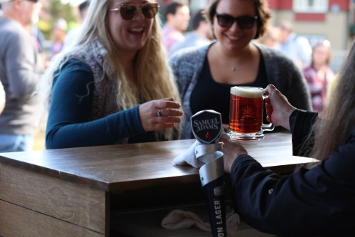 Two women smile at a bar while one holds a beer mug, with a Sam Adams tap in the foreground.