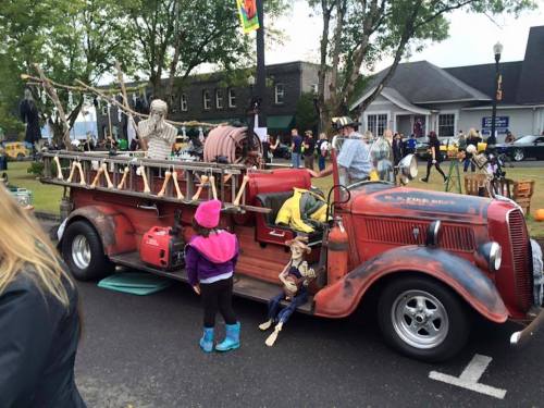 A vintage fire truck decorated with puppets and props, with a child in a pink hat admiring it.