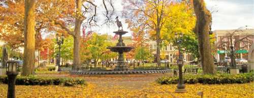 A scenic park in autumn, featuring a fountain surrounded by colorful trees and fallen leaves.