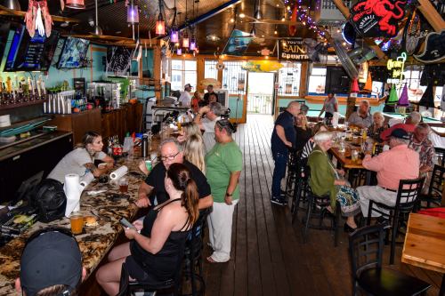A lively bar scene with patrons at the counter and tables, colorful decor, and a bartender serving drinks.