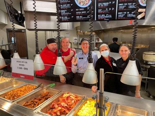 A group of five restaurant staff members smiling behind a food counter with various dishes and a menu overhead.