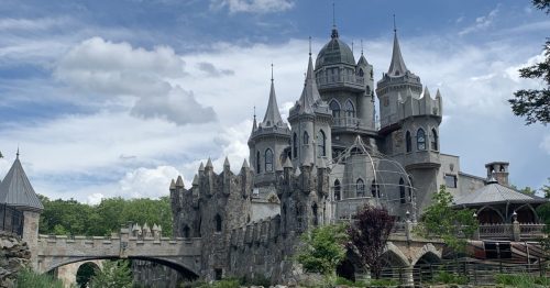 A majestic castle with turrets and a stone bridge, reflected in a calm body of water under a cloudy sky.