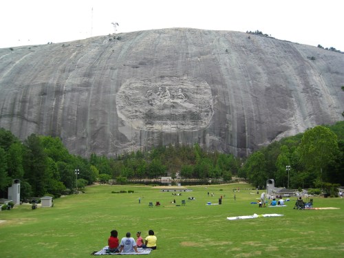 A large granite mountain with a carved relief, surrounded by green grass and people enjoying the park below.