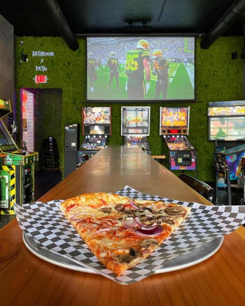 A slice of pizza on a checkered plate in a vibrant bar with arcade games and a sports screen in the background.