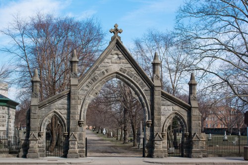 Gothic stone archway with a cross, leading into a tree-lined cemetery under a clear blue sky.