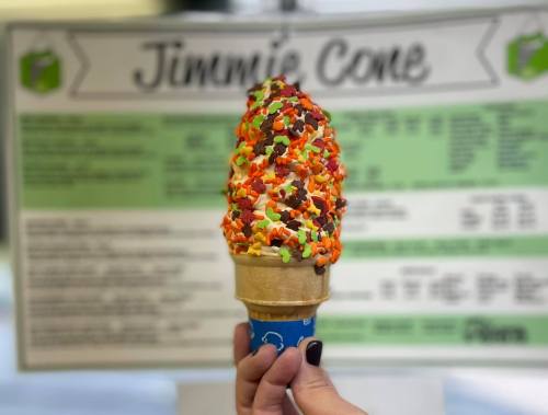 A hand holding a colorful ice cream cone topped with sprinkles, in front of a menu board.