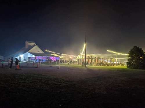 A nighttime outdoor event with illuminated tents and a building, surrounded by people and string lights.