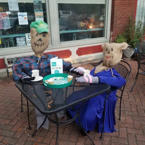 Two scarecrows sit at a table outside, enjoying tea, dressed in casual and formal attire.