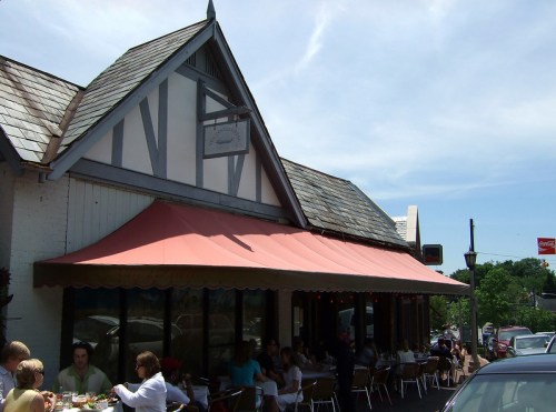 A charming restaurant with a sloped roof and outdoor seating, bustling with diners on a sunny day.
