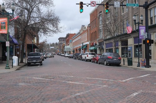 A brick-paved street lined with shops and parked cars, featuring traffic lights and American flags on buildings.