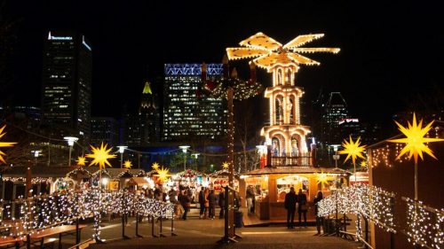 A festive night scene of a Christmas market with twinkling lights, a tall illuminated structure, and city skyline in the background.