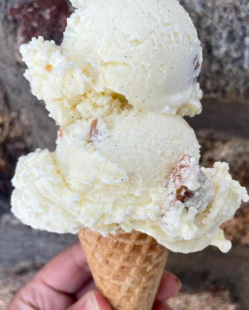 A hand holding a waffle cone with two scoops of creamy vanilla ice cream, featuring bits of nuts.