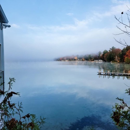 A serene lake scene with fog, reflecting trees and a dock, under a clear blue sky.