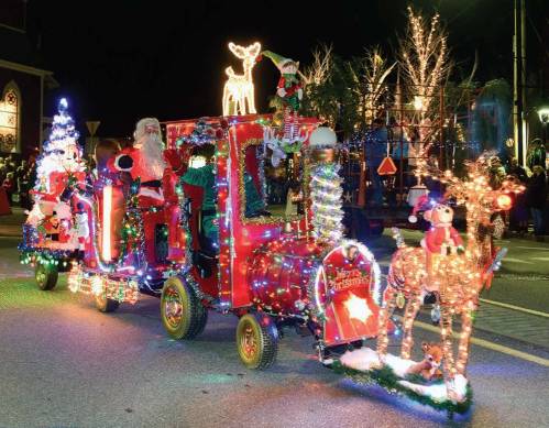 A festive parade float featuring Santa, a decorated train, and illuminated reindeer, surrounded by holiday lights.