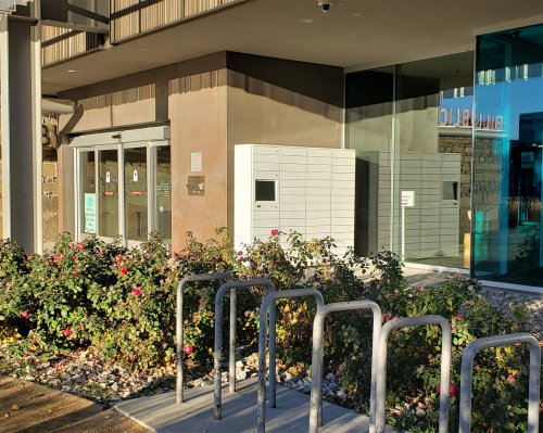 A modern building entrance with a package locker and bike rack, surrounded by flowering bushes.