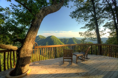 A wooden deck with two chairs overlooks lush green mountains under a clear blue sky.