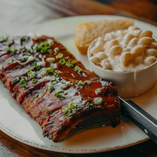 A plate featuring glazed ribs topped with green onions, a side of macaroni and cheese, and a cornbread muffin.