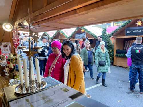 Two women smile while looking at items in a festive market, surrounded by holiday decorations and other shoppers.