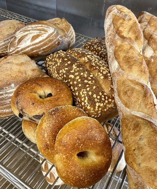 A variety of freshly baked breads, including bagels, loaves with sesame seeds, and baguettes, on a wire rack.