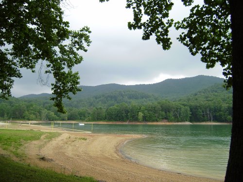A serene lake surrounded by green trees and mountains under a cloudy sky, with a sandy beach in the foreground.