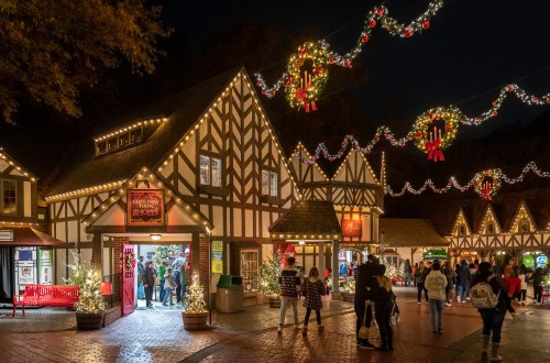 A festive village scene at night, adorned with Christmas lights, wreaths, and people enjoying the holiday atmosphere.