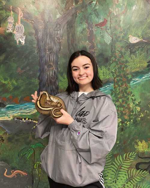 A young woman smiles while holding a snake in front of a colorful, nature-themed mural.