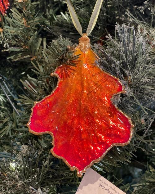 A vibrant red and orange leaf-shaped ornament hanging on a Christmas tree, surrounded by green pine needles.
