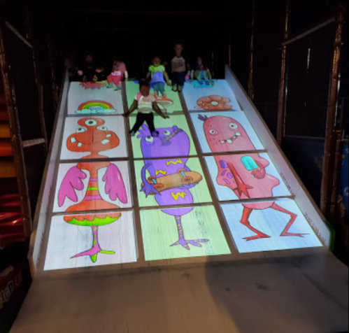 Children play on a colorful slide featuring cartoon monster illustrations projected on the surface.