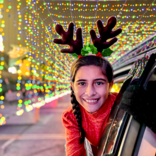 A girl with reindeer antlers smiles from a car, surrounded by colorful holiday lights.