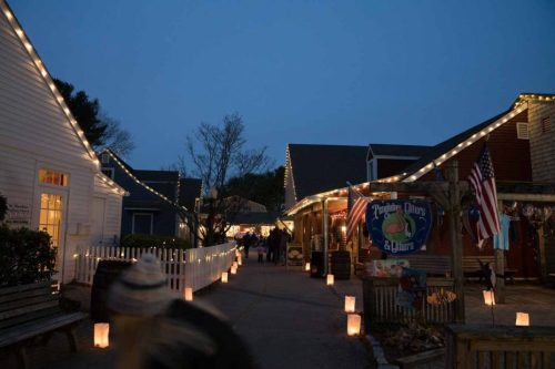 A charming street lined with shops and glowing lanterns, illuminated by string lights at dusk.