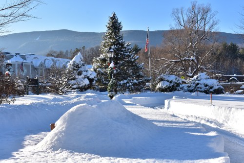A snowy landscape with a tree, lamp post, and mountains in the background under a clear blue sky.