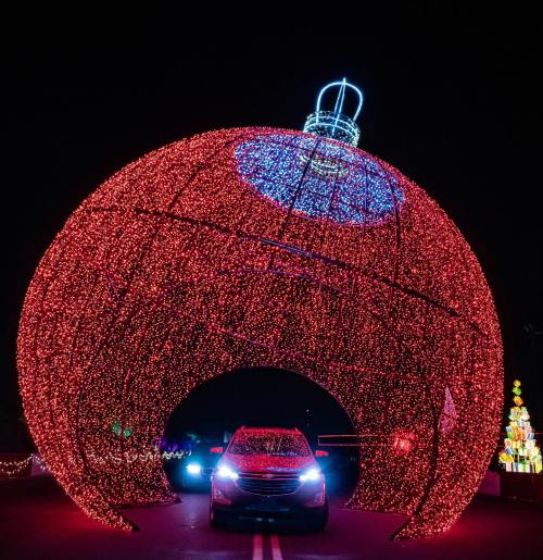 A car drives through a large, illuminated Christmas ornament made of red lights at night.