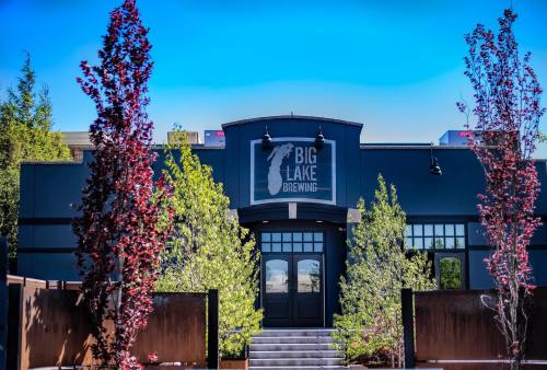 Exterior of Big Lake Brewing, featuring a dark building with a logo, surrounded by trees and a clear blue sky.