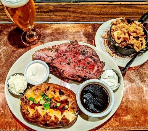 A plate with prime rib, baked potato, dipping sauces, and a side of crispy onions, accompanied by a glass of beer.