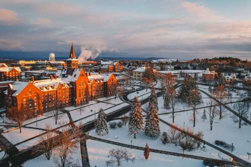 A snowy campus scene at sunset, featuring historic buildings, trees, and a clock tower against a colorful sky.