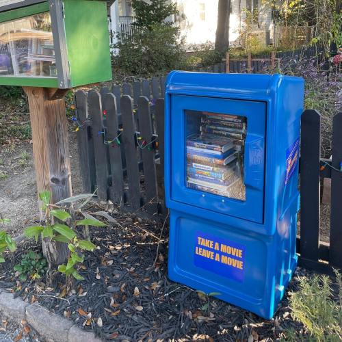 A blue movie exchange box next to a green mailbox, surrounded by greenery and a wooden fence.