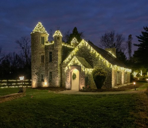 A charming stone house adorned with festive lights, set against a twilight sky and surrounded by greenery.