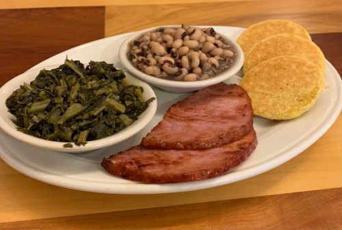 A plate featuring two slices of ham, black-eyed peas, collard greens, and three cornbread cakes.
