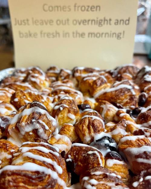 A close-up of a baked pastry topped with icing, with a sign in the background about overnight preparation.